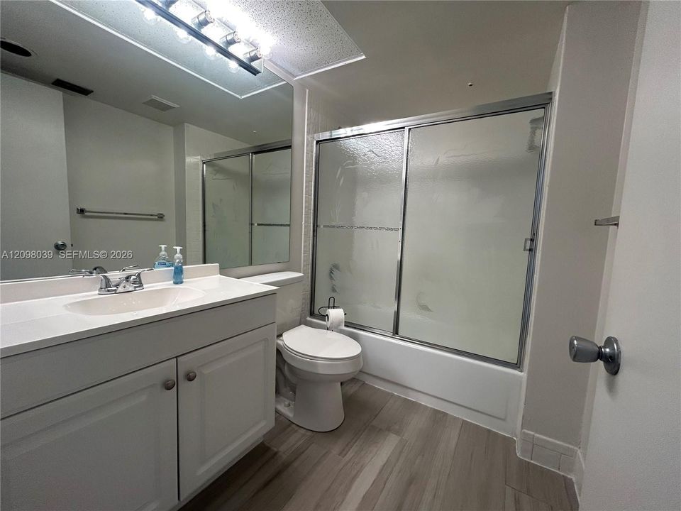 Bathroom featuring a white vanity cabinet with an integrated sink, a large wall-mounted mirror, a tub-shower combination with a sliding glass enclosure, and wood-finish flooring