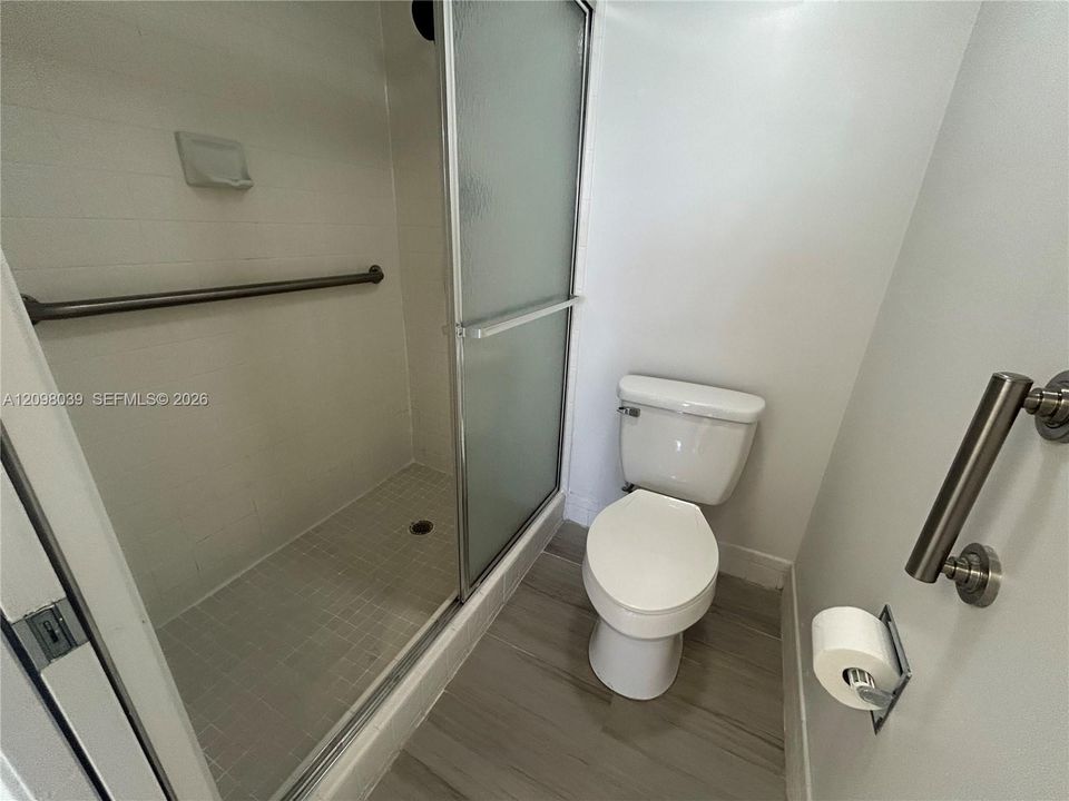 Walk-in shower with a frosted glass sliding door, tiled surround, and integrated wall-mounted grab bars, accompanied by a standard-height toilet and wood-finish flooring