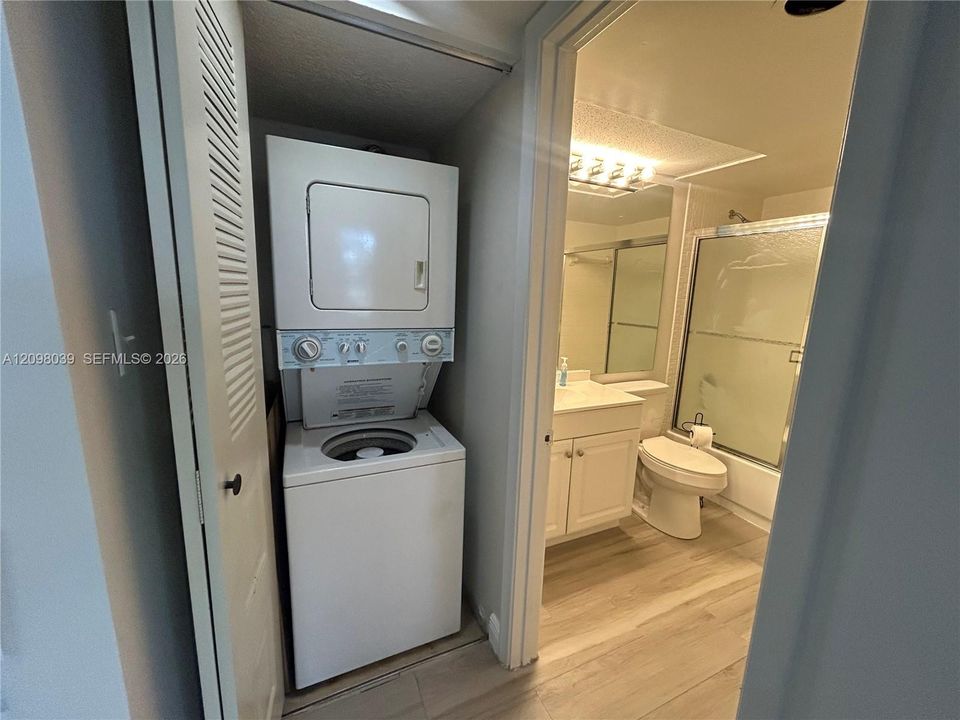 Stacked laundry unit set within a louvered closet, wood-finish flooring, white vanity with integrated basin, and a tub-shower combination with a glass enclosure