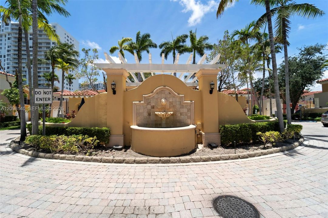 Custom stone-finish fountain featuring an integrated wall feature, overhead pergola, symmetrical wall-mounted light fixtures, and paver stone circular driveway