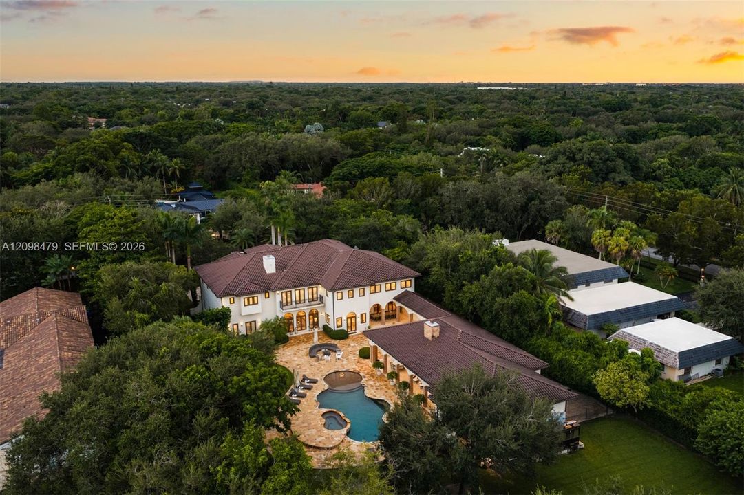 Aerial perspective of an expansive residence featuring a multi-gabled tile roof, stone-paved pool deck, custom-shaped swimming pool with integrated spa, and mature perimeter landscaping