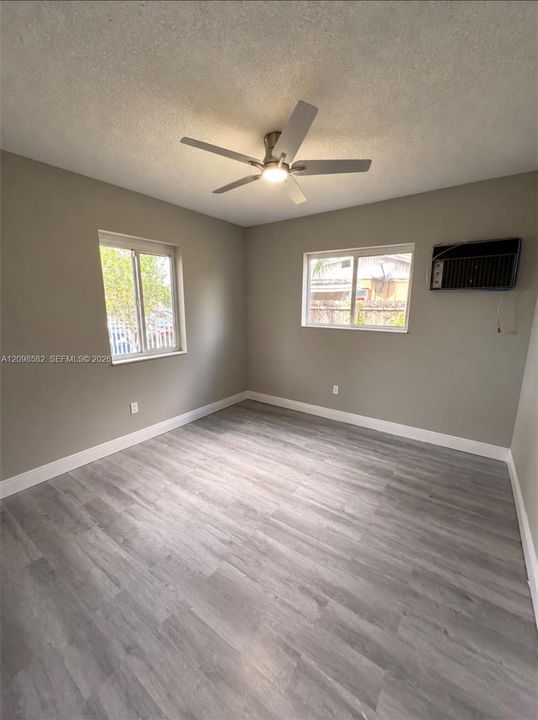 Room featuring wood-finish flooring, dual windows, a ceiling fan with integrated lighting, and a wall-mounted air conditioning unit