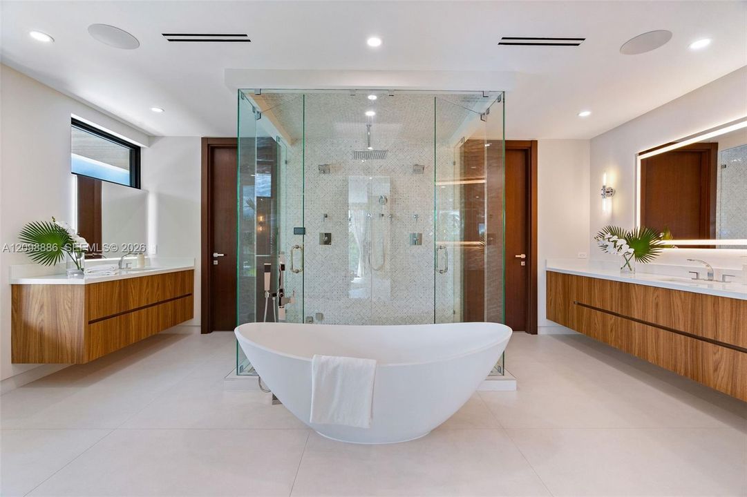 Dual floating wood-finish vanities with rectangular vessel sinks, floor-to-ceiling glass-enclosed shower with tiled interior, and freestanding soaking tub