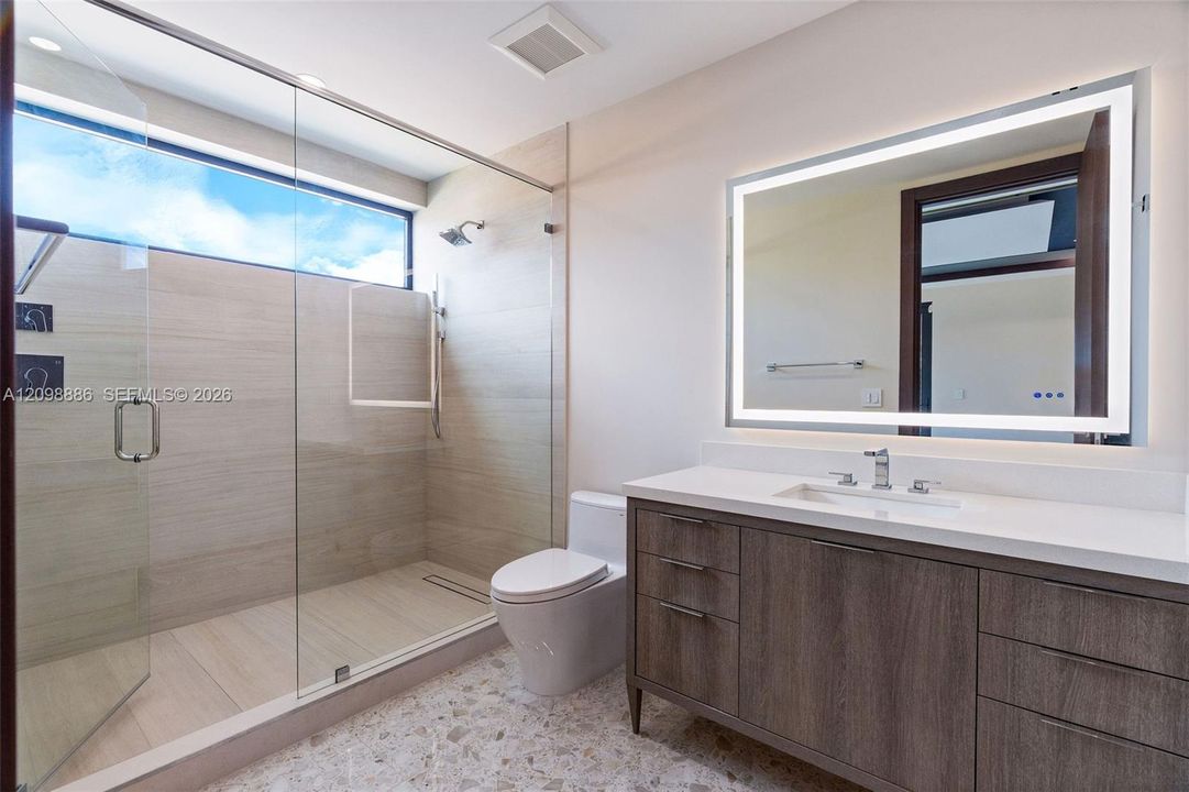 Bathroom featuring a walk-in shower with large-format tile surround, frameless glass enclosure, a wood-finish vanity with a stone countertop, and an integrated LED-lit vanity mirror