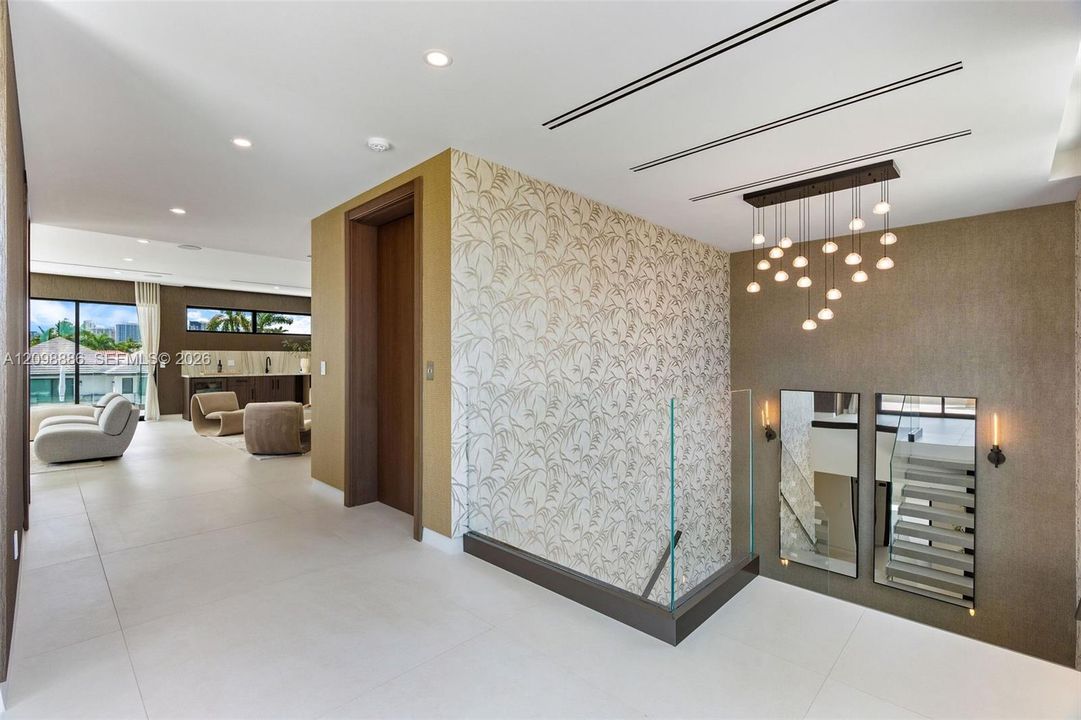 Interior space featuring large-format tile flooring, patterned accent wall, glass panel railing, multi-pendant chandelier, and recessed linear lighting