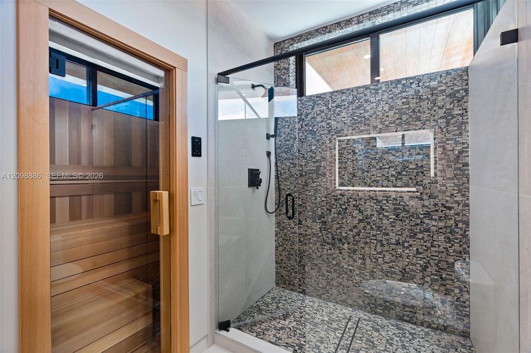Built-in wood-paneled sauna featuring a glass-door entrance, accompanied by a walk-in shower with a mosaic-tiled accent wall, glass enclosure, transom window, and matte black fixtures