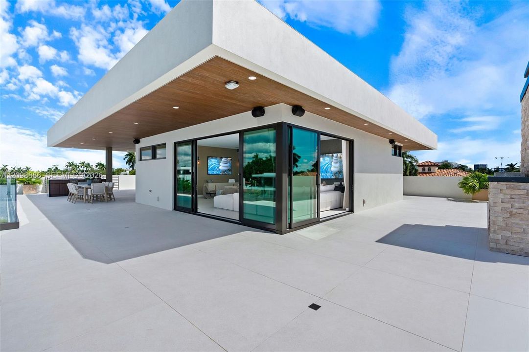 Expansive rooftop terrace featuring large-format tile flooring, a cantilevered roof overhang with wood-finish soffits, recessed lighting, and floor-to-ceiling multi-panel sliding glass doors