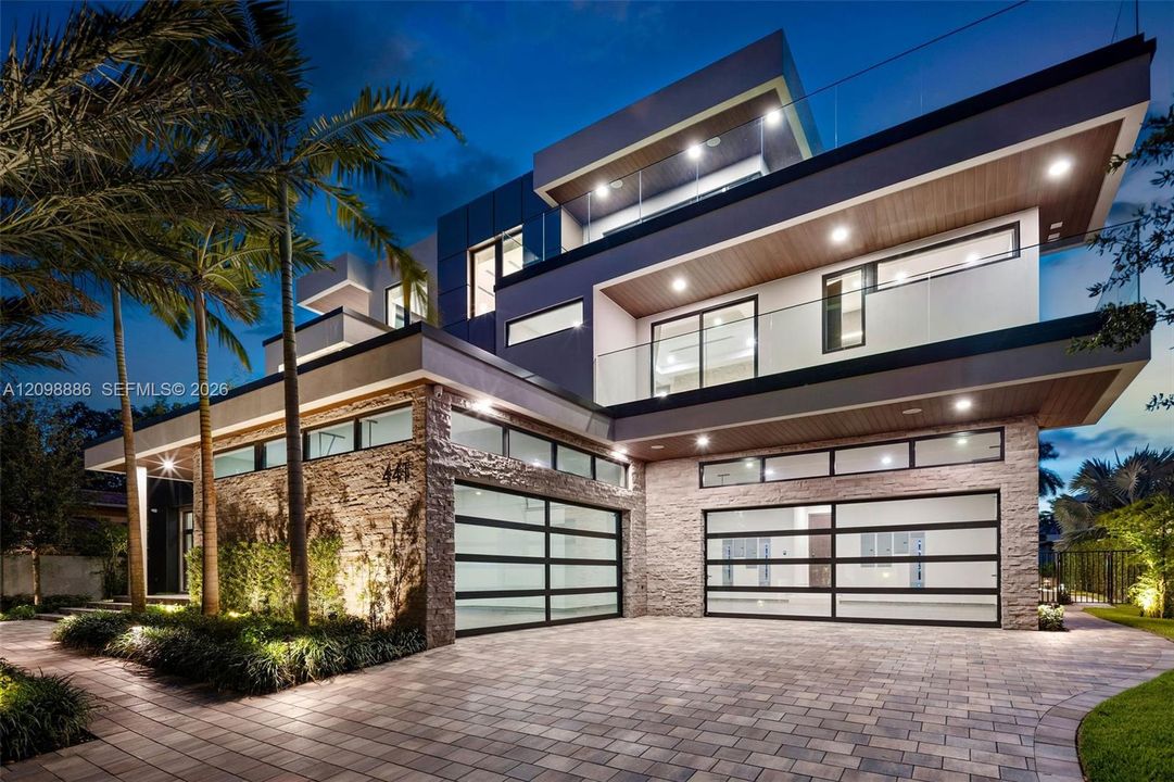 Contemporary multi-level exterior featuring stacked stone masonry, horizontal glass panel garage doors, glass-railed balconies, and wood-finish soffits