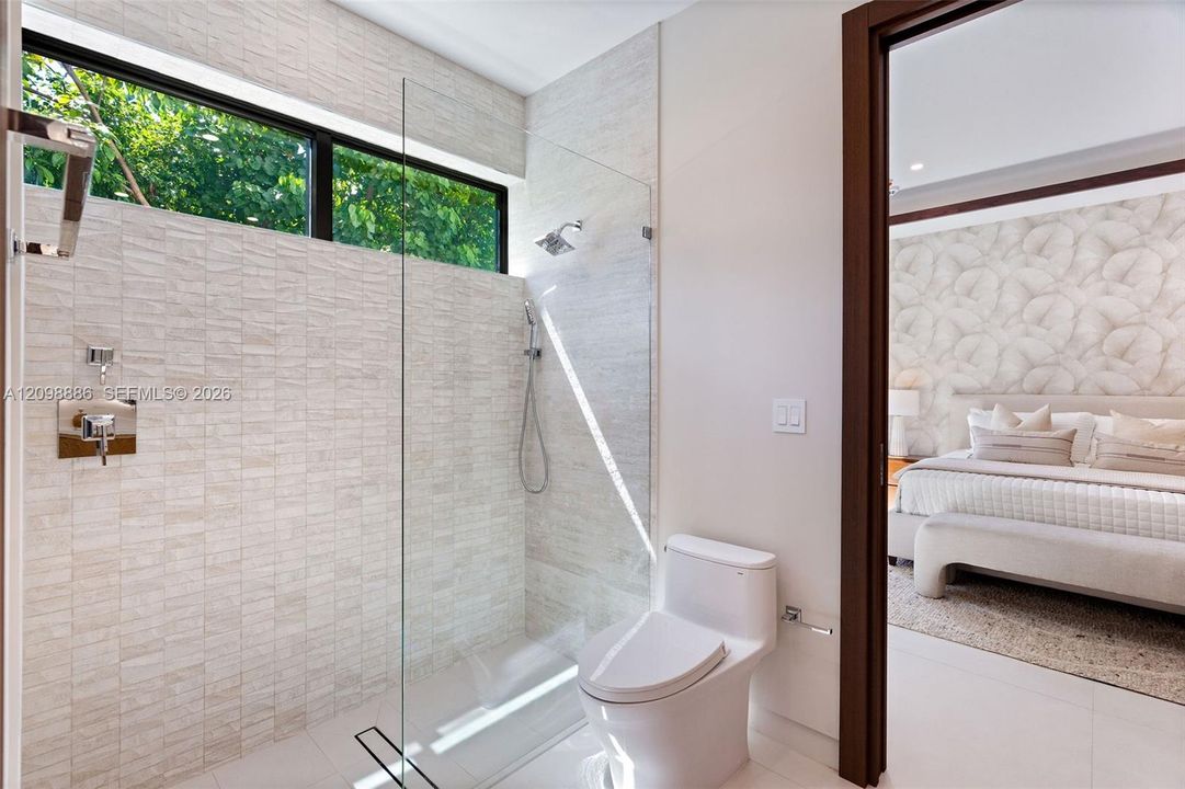 Walk-in shower featuring textured stone tile walls, a frameless glass panel, a rainfall showerhead, and a rectangular window, complemented by a one-piece elongated toilet