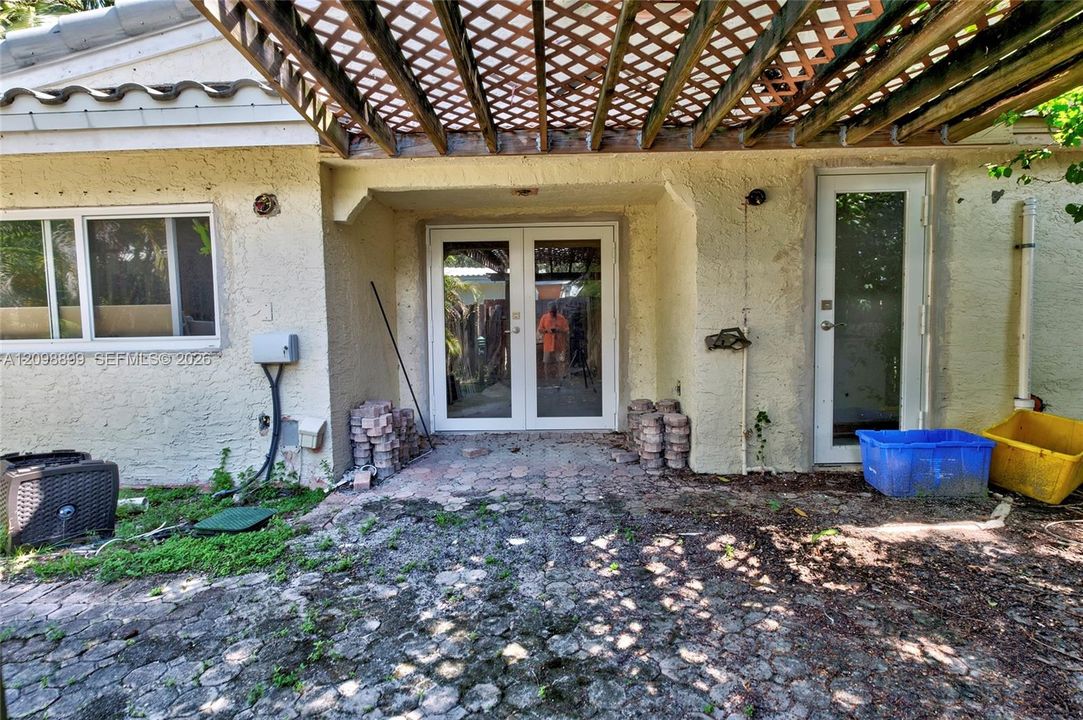Exterior patio space featuring a latticed wood-frame pergola, stone paver flooring, double-glass entry doors, and a secondary single-glass door