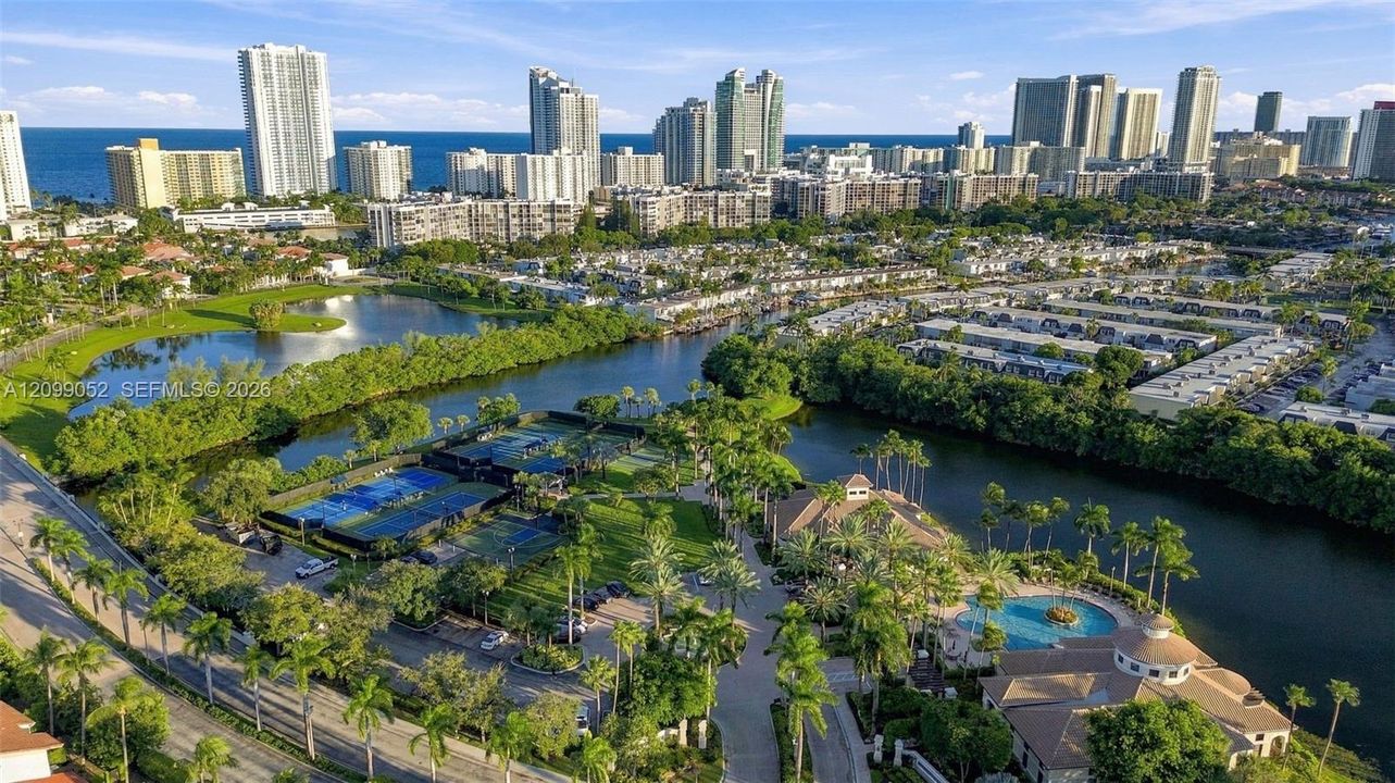 Aerial perspective of the community featuring a swimming pool, multiple tennis courts, a central lagoon, and landscaped grounds with mature palm trees
