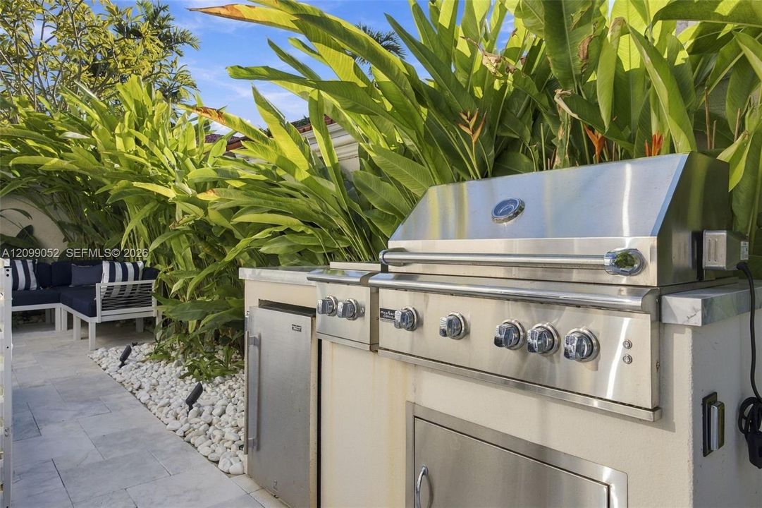 Outdoor kitchen featuring a stainless steel grill, stone-surfaced countertops, custom built-in cabinetry, and integrated storage, set against a landscaped garden bed