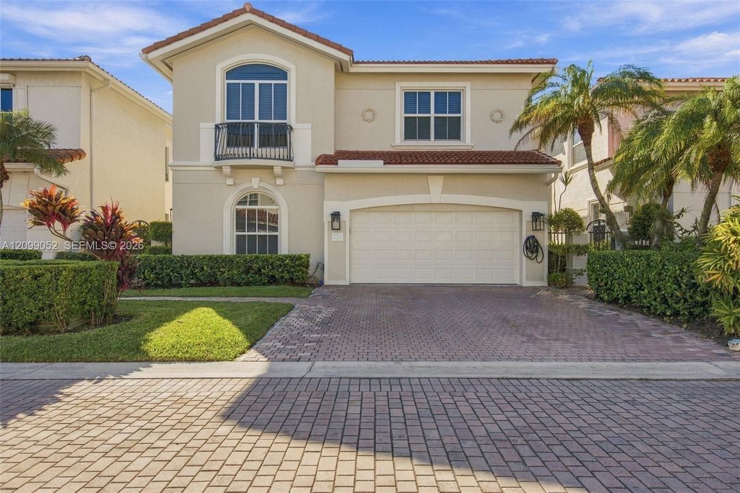 Two-story stucco exterior featuring a tile roof, an arched balcony with metal railing, and a paver driveway leading to a two-car garage