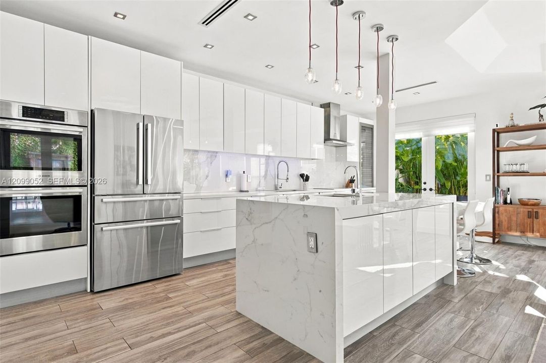 Modern kitchen featuring high-gloss white cabinetry, a center island with a stone-finish waterfall countertop, stainless steel double wall ovens, wood-finish flooring, and pendant lighting