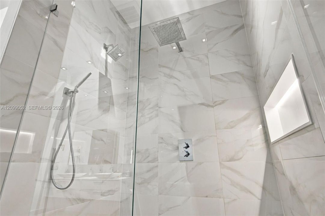 Walk-in shower featuring large-format light veined tile, frameless glass enclosure, ceiling-mounted rainfall showerhead, adjustable handheld shower wand, and integrated recessed shelving with integrated lighting