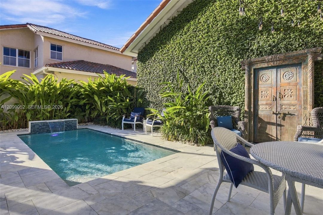 Outdoor space featuring a swimming pool with a built-in water feature, stone paver decking, decorative wood-carved gate, and exterior wall with climbing vegetation