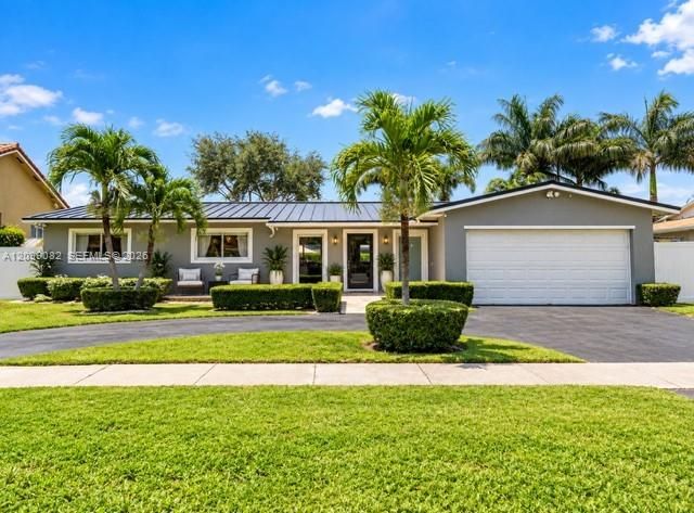 Single-story residence featuring a standing-seam metal roof, attached two-car garage, circular asphalt driveway, and manicured landscaping with mature palms