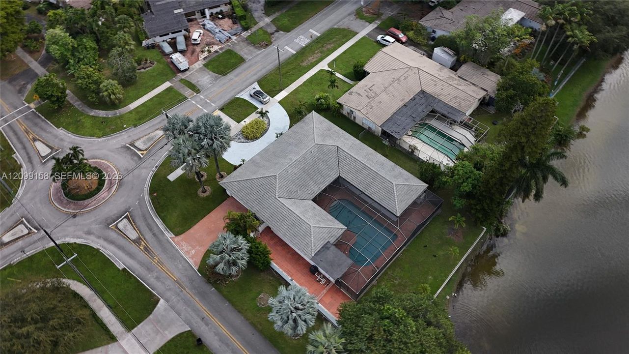 Aerial perspective of a corner-lot property featuring a tile roof, an enclosed swimming pool, brick-paved driveway, and water frontage
