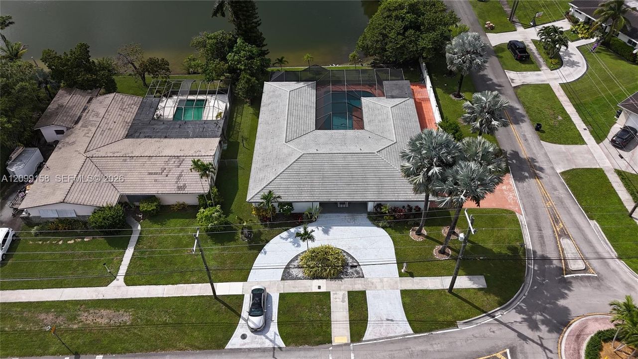 Aerial perspective of waterfront property featuring a U-shaped home, screened-in pool enclosure, circular concrete driveway, and mature landscaping