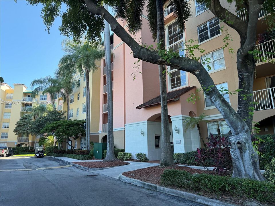 Multi-story exterior featuring a stucco facade, multi-level balconies with white railings, and an arched entry portico, surrounded by mature palms and landscaping