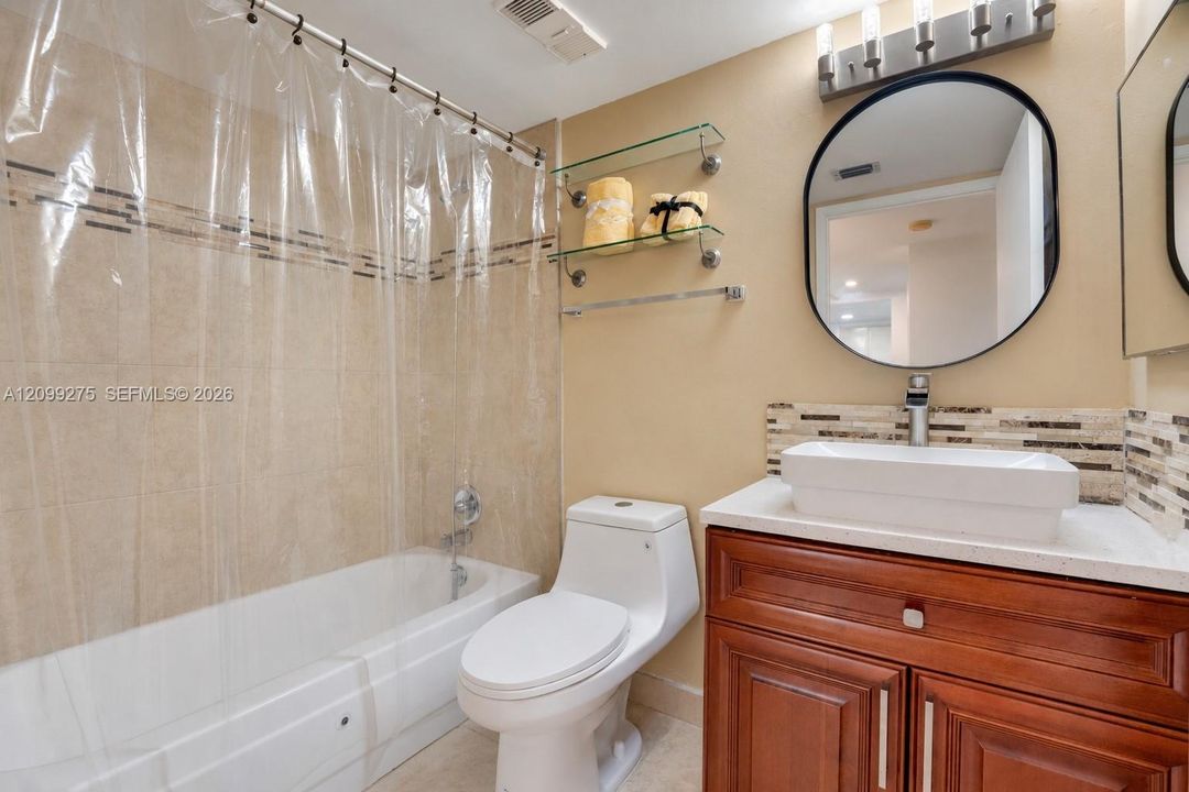 Well-appointed bathroom featuring a modern vessel sink, wood vanity, large round mirror, and a bathtub with tiled walls and a clear shower curtain.