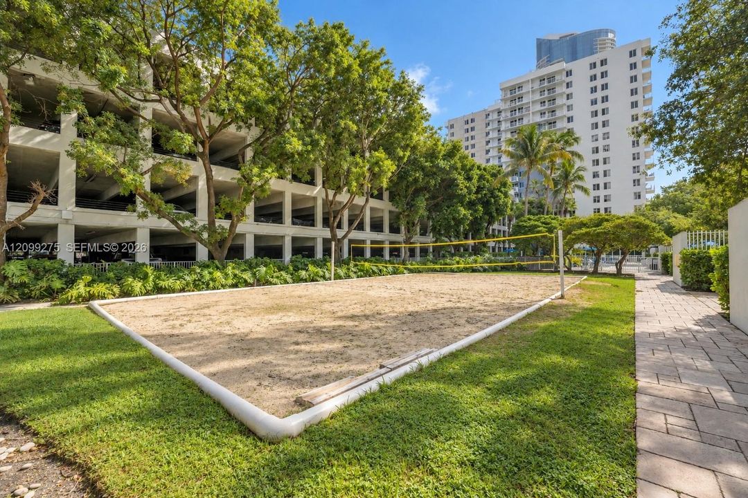 Outdoor sand volleyball court surrounded by lush tropical landscaping, mature shade trees, and residential high-rises, offering residents a relaxing space for recreation and outdoor activities.