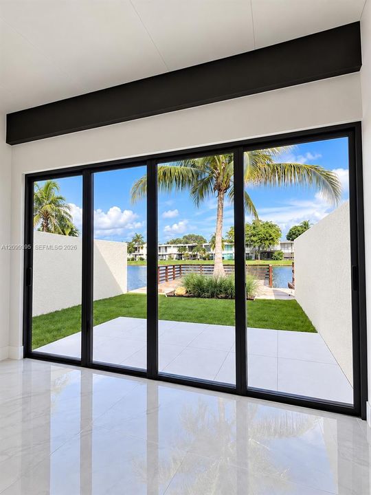 Floor-to-ceiling multi-panel sliding glass doors with matte black framing, exposed structural ceiling beam, polished stone-finish flooring, paved exterior patio, and water frontage