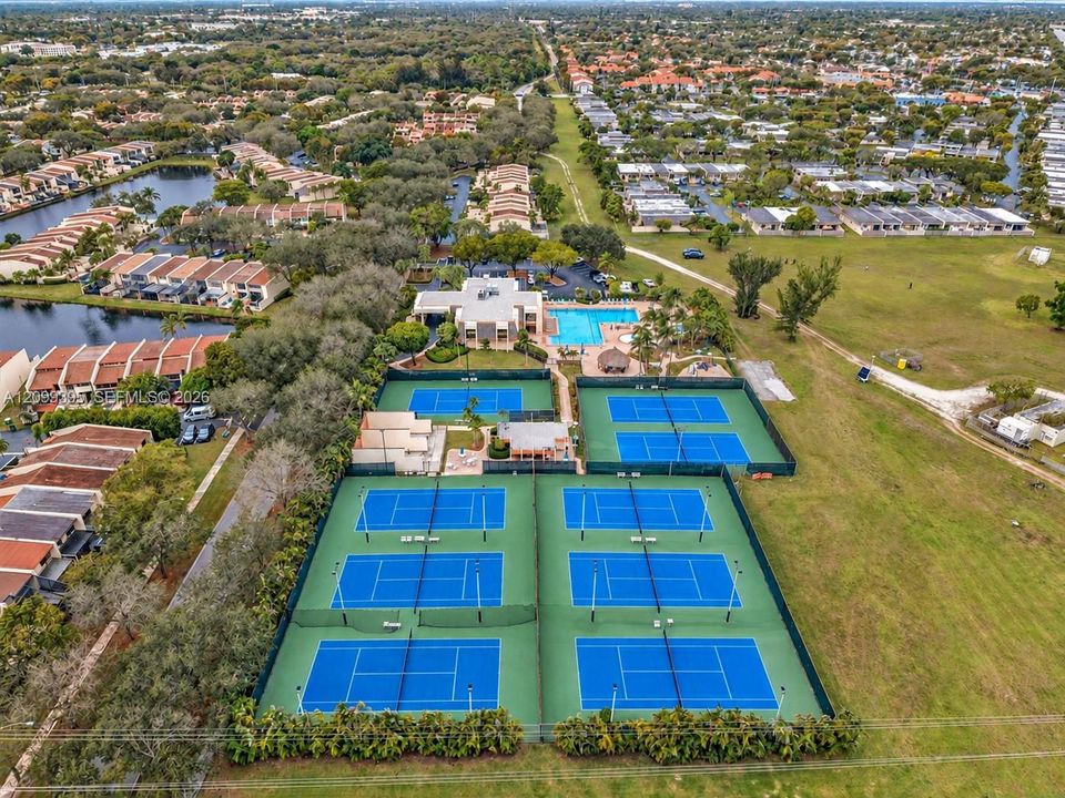 Aerial perspective of a multi-court tennis facility featuring six fenced hard courts, a swimming pool, and a clubhouse