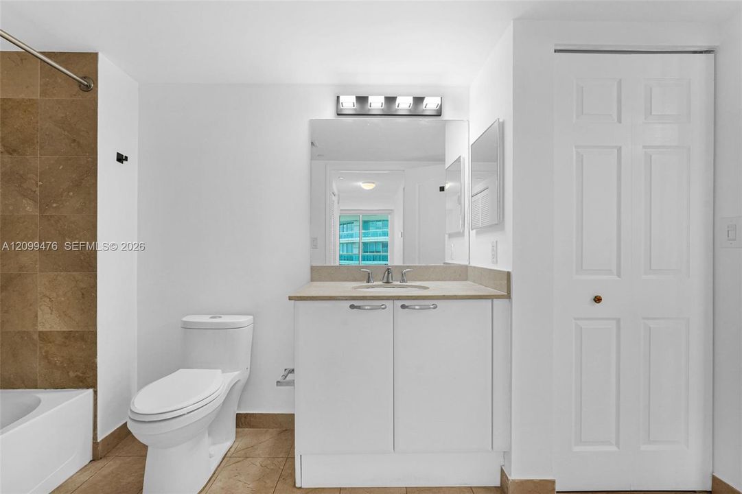 Bathroom featuring a vanity with neutral stone-finish countertop, undermount sink, multi-bulb wall-mounted light fixture, large vanity mirror, and bathtub surround with neutral tile finish