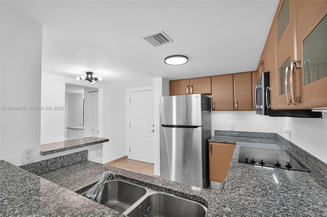 Kitchen featuring stone countertops, wood-finish cabinetry with glass-paneled uppers, stainless steel refrigerator, and dual-basin stainless steel sink with high-arc faucet