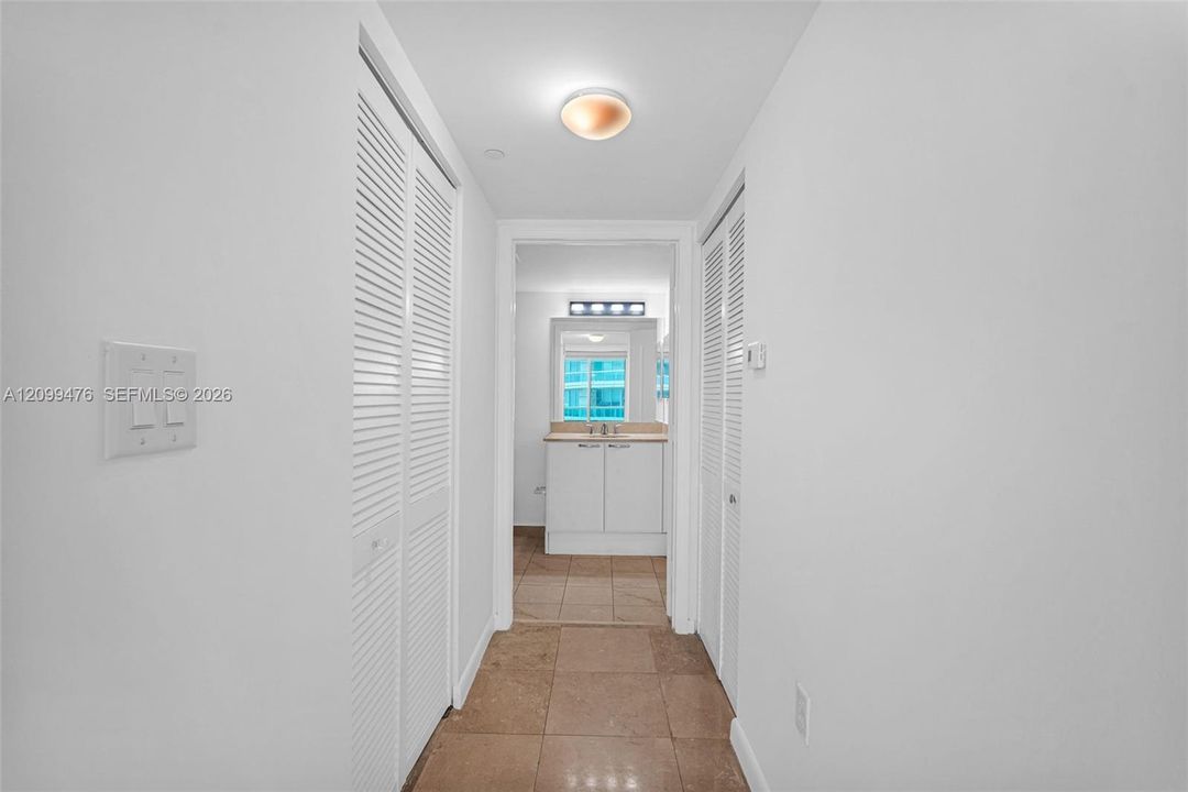 Hallway featuring dual sets of white louvered-door closets, neutral-toned tile flooring, and a flush-mount ceiling light fixture