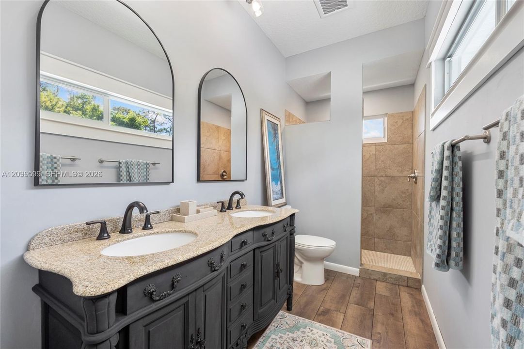 Dual-sink vanity with stone countertop and dark cabinetry, arch-top mirrors, wood-finish flooring, and a tiled shower enclosure