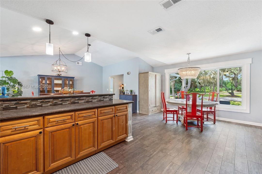 Kitchen featuring wood cabinetry, stone-finish countertops with a mosaic tile backsplash, wood-finish flooring, vaulted ceilings, and large windows with integrated bead-embellished pendant lighting