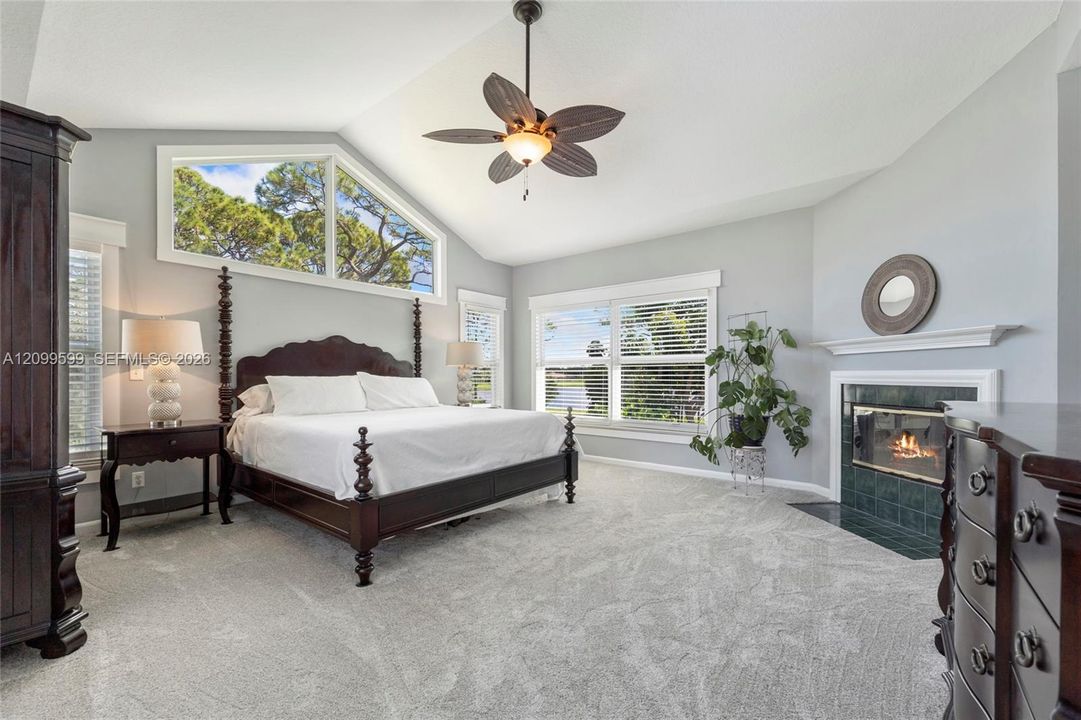 Vaulted ceiling with a central multi-blade ceiling fan, expansive trapezoidal window, fireplace with a tiled hearth and mantel, and wall-to-wall carpeting