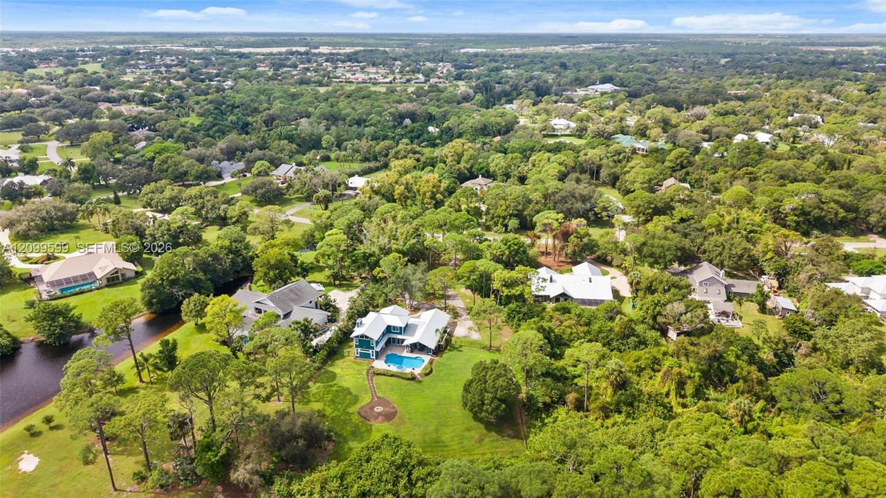 Aerial perspective featuring a multi-story residence with a metal roof, an in-ground swimming pool, a stone-paved patio, and established mature trees on the grounds