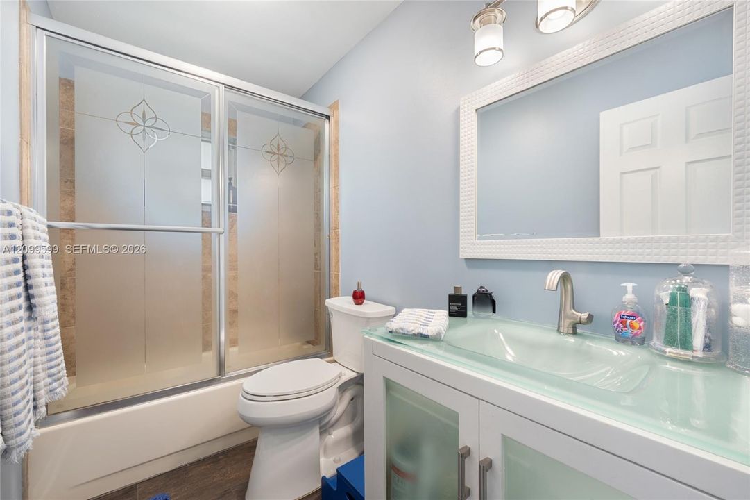 Bathroom featuring a vanity with an integrated glass sink and countertop, frosted glass cabinet inserts, a shower enclosure with decorative etched glass doors, a wall-mounted framed mirror, and wood-finish flooring