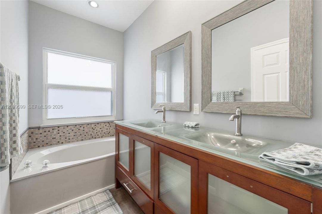 Dual-sink vanity with a translucent glass countertop and wood cabinetry, paired with a built-in soaking tub featuring mosaic tile surround and dual wall-mounted mirrors