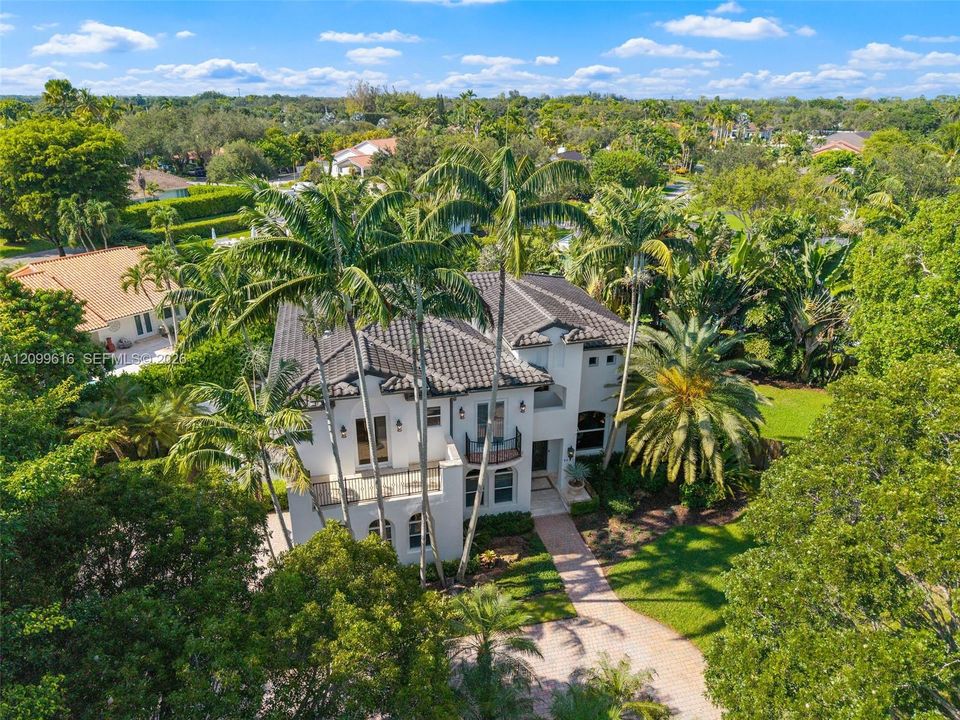 Two-story estate with light-toned stucco exterior, multi-level balconies, decorative iron railings, and a tiled roof, set on a landscaped lot with mature palm trees and a paved driveway