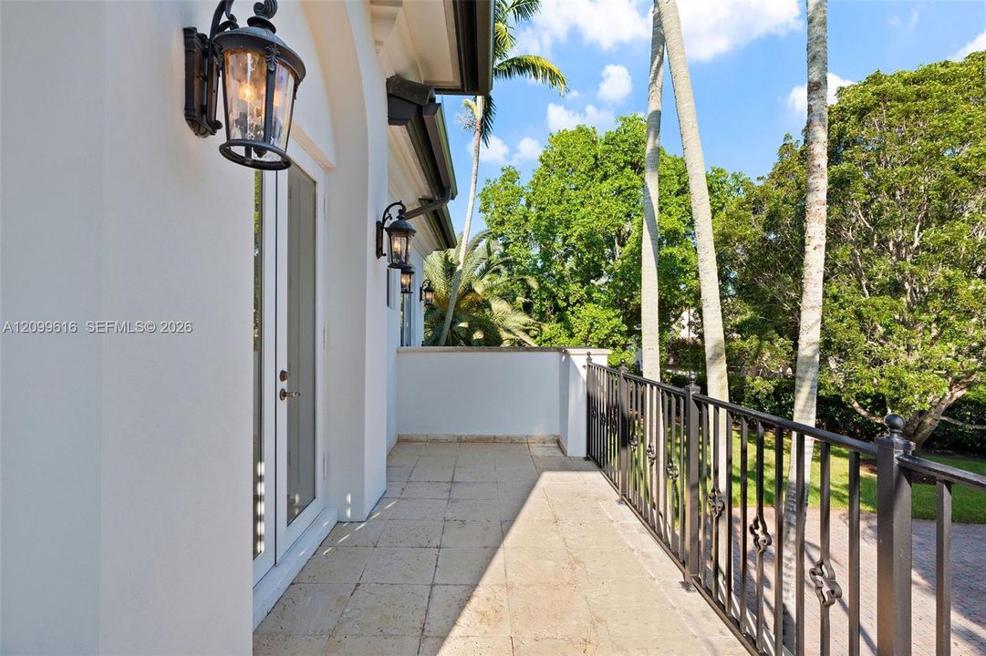 Covered balcony featuring stone-finish tile flooring, decorative metal railings, and glass-paneled double doors accented by wall-mounted coach lighting