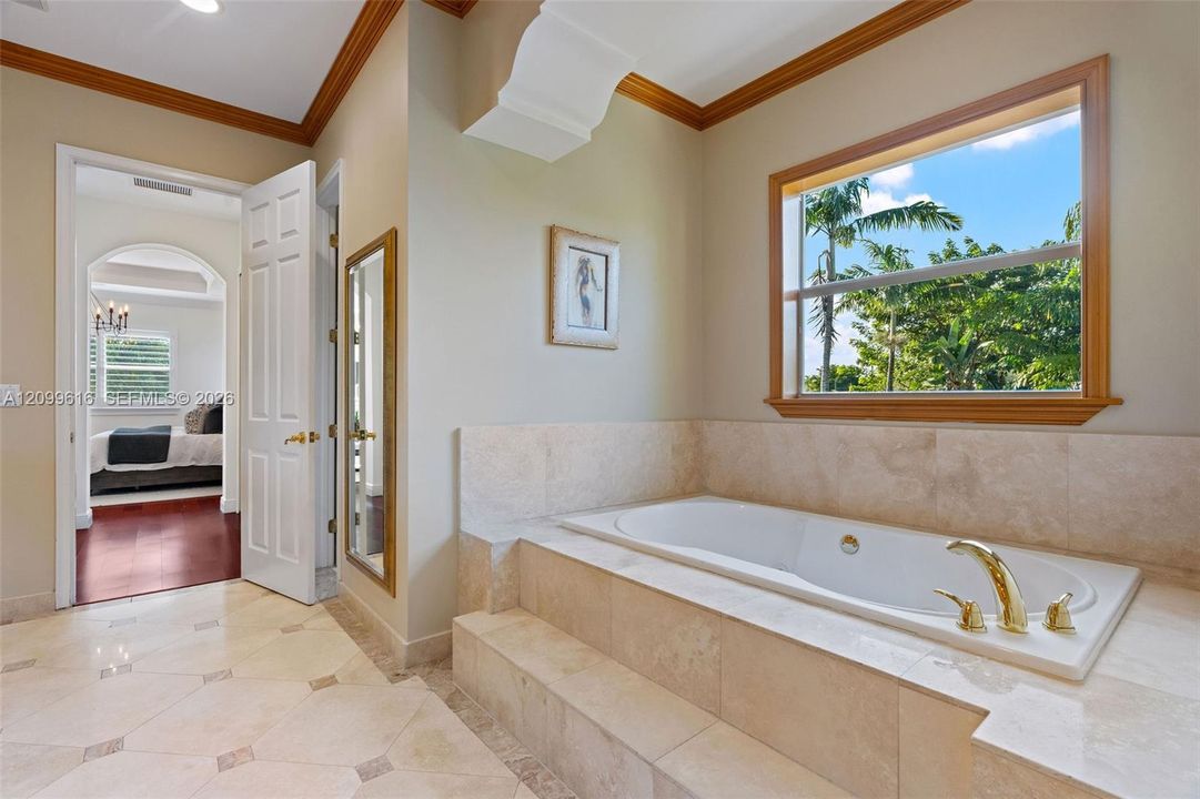 Soaking tub with gold-tone hardware, stone tile surround and flooring, wooden window casing, and crown molding