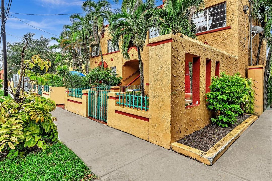 Stucco exterior featuring red trim, decorative metal fencing, mature palm trees, and established landscaping