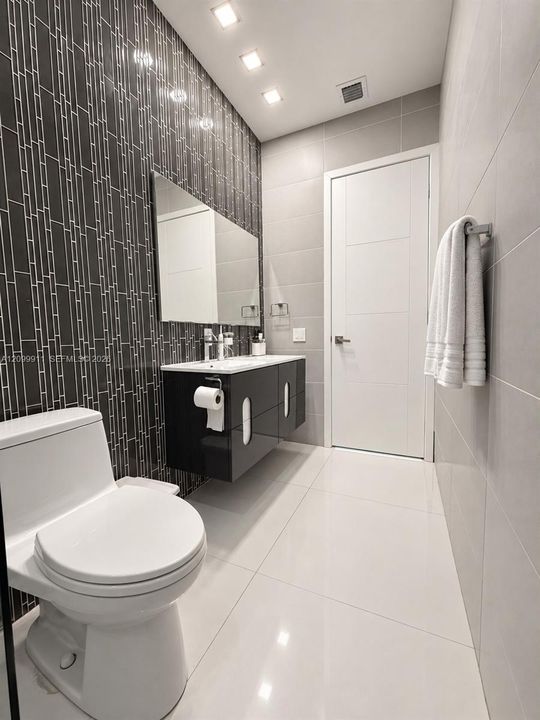 Bathroom featuring large-format white tile flooring, wall-mounted black cabinetry, a vertical-patterned dark tile accent wall, recessed lighting, and a frameless mirror