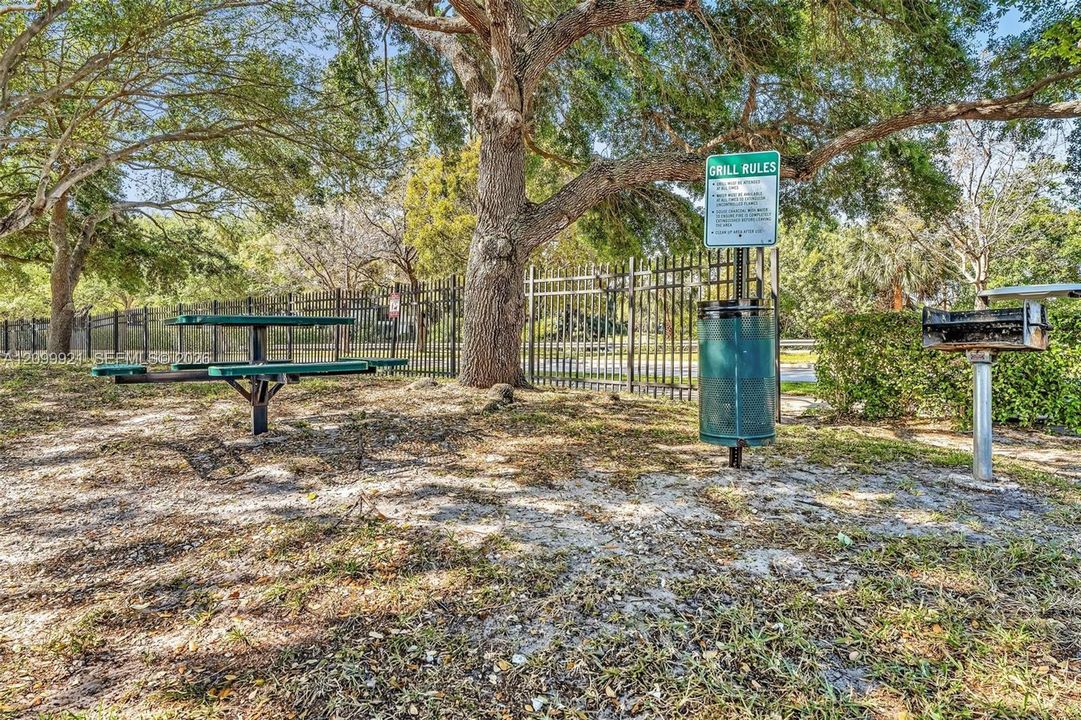 Outdoor recreational area featuring a metal picnic table, pedestal-mounted grill, waste receptacle, and mature canopy trees enclosed by perimeter metal fencing