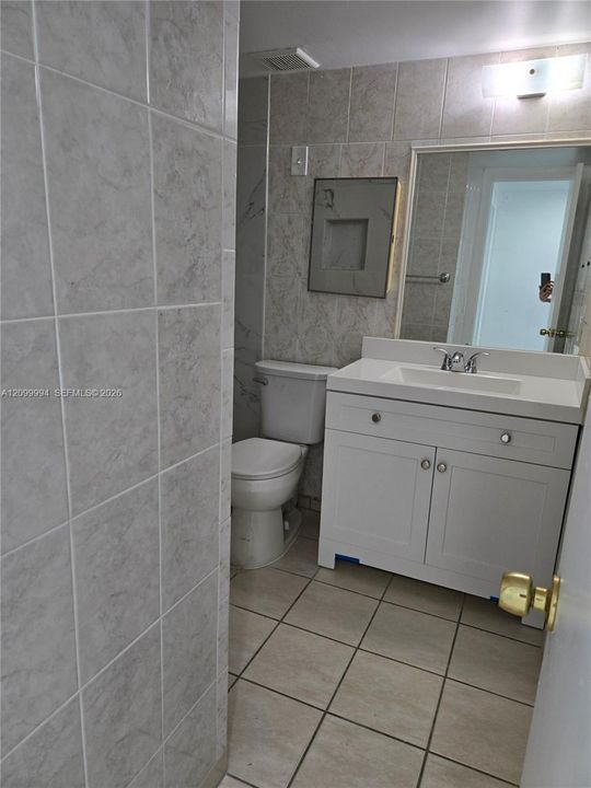 Bathroom featuring a white vanity cabinet with a stone-top integrated sink, neutral wall-to-wall ceramic tile, recessed shower niche, and a wall-mounted rectangular mirror
