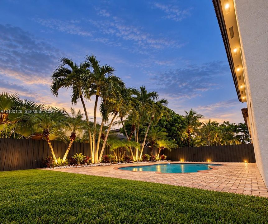 Private outdoor space featuring a swimming pool, stone-paver patio, manicured lawn, perimeter fencing, and mature palm trees