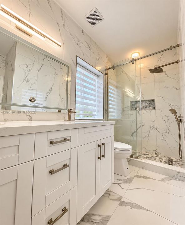 White vanity with stone-finish countertop, floor-to-ceiling stone-finish wall tile, walk-in glass-enclosed shower featuring a rainfall showerhead, and integrated wall niche