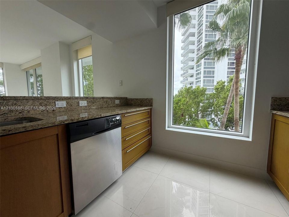 Kitchen featuring natural stone countertops, warm-toned wood cabinetry, stainless steel dishwasher, floor-to-ceiling windows, and polished tile flooring