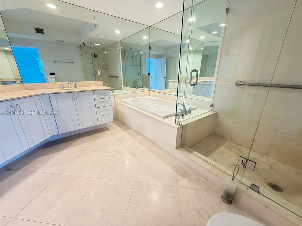 Bathroom featuring a double vanity with light-toned countertop, soaking tub with tile surround, glass-enclosed shower, expansive wall-to-wall mirrors, and stone-finish tile flooring