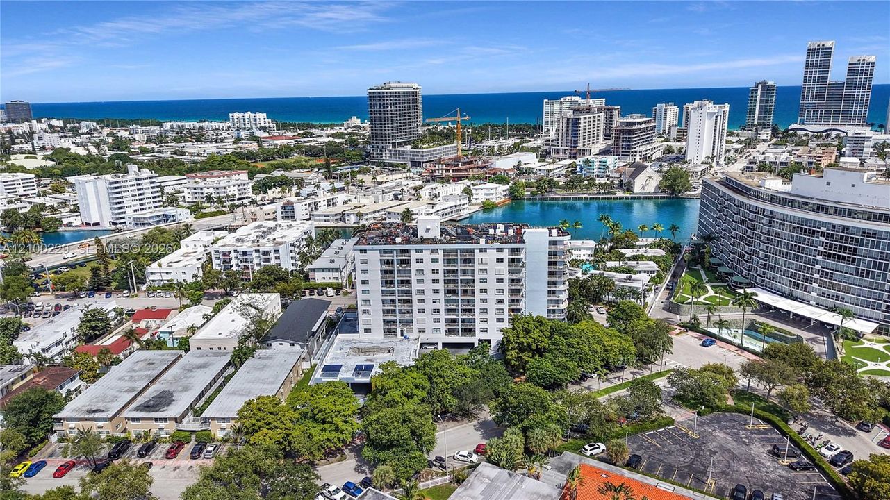 Aerial perspective of a high-rise condominium complex featuring a coastal location with shoreline proximity, mature tree cover, paved parking, and a mid-rise structural design
