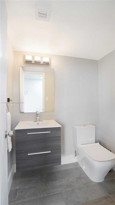 Wall-mounted dark wood-finish vanity with a white rectangular countertop basin, polished chrome faucet, rectangular frameless mirror, and neutral-toned stone-finish tile flooring
